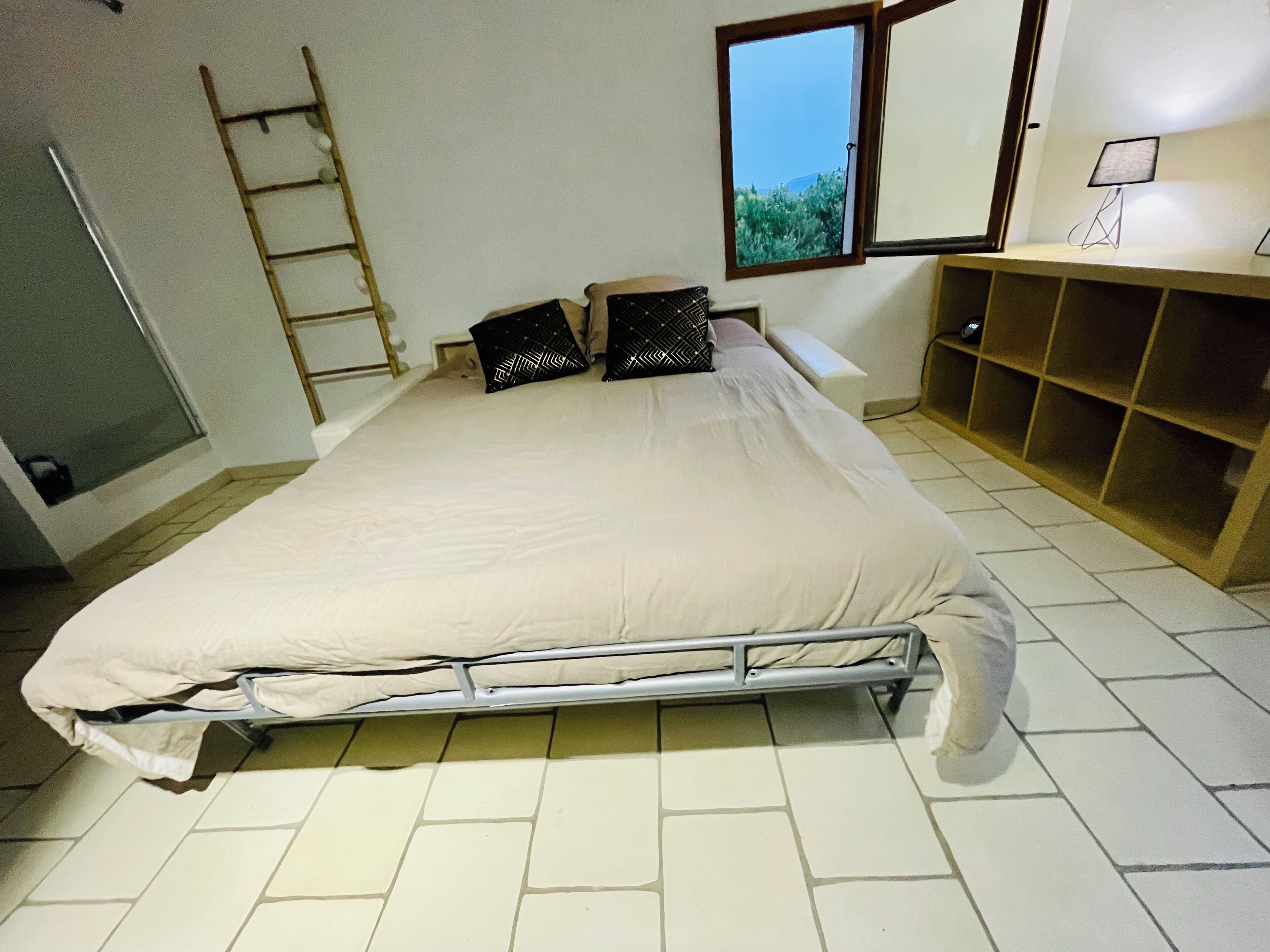 A bedroom with a bed, pillows, a ladder, a window, and a desk with a lamp.