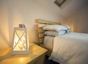 A neatly made bed with white bedding and striped pillows, a wooden nightstand with a white lantern, and a wooden headboard. - Saltee View Holiday Home (Kilmore)