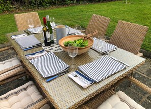 A wicker dining table set outdoors with striped placemats, wine glasses, and a bowl of greens. - Saltee View Holiday Home (Kilmore)