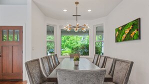 A dining area with a long table, six chairs, a chandelier, and a large window with a view of greenery.