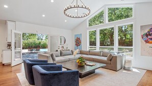 A spacious living room with a large sofa, two armchairs, and a coffee table. There are large windows providing a view of the outdoors.