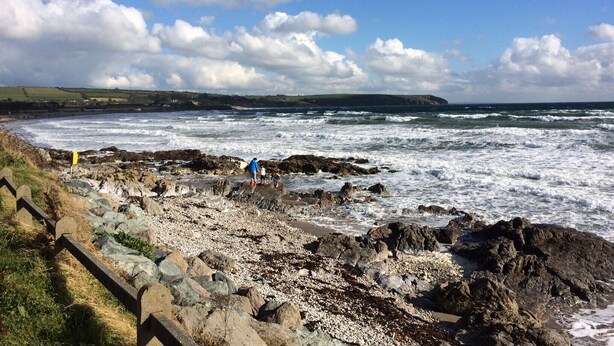 A rocky coastal area with waves crashing onto the shore, a clear sky, and a distant landmass.