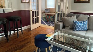 Un salon avec une table basse en verre, un canapé agrémenté de coussins à motifs, un pouf bleu et un plancher en bois.