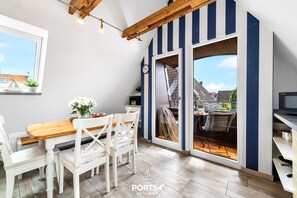 Un coin repas avec une table en bois et des chaises blanches, une horloge au mur et une vue sur l’extérieur à travers une porte vitrée.