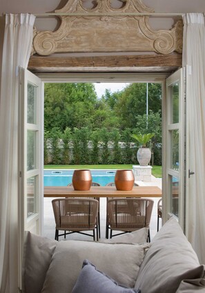 A room with a view of a swimming pool, two wicker chairs, a table with two copper vases, and a cushioned bench.