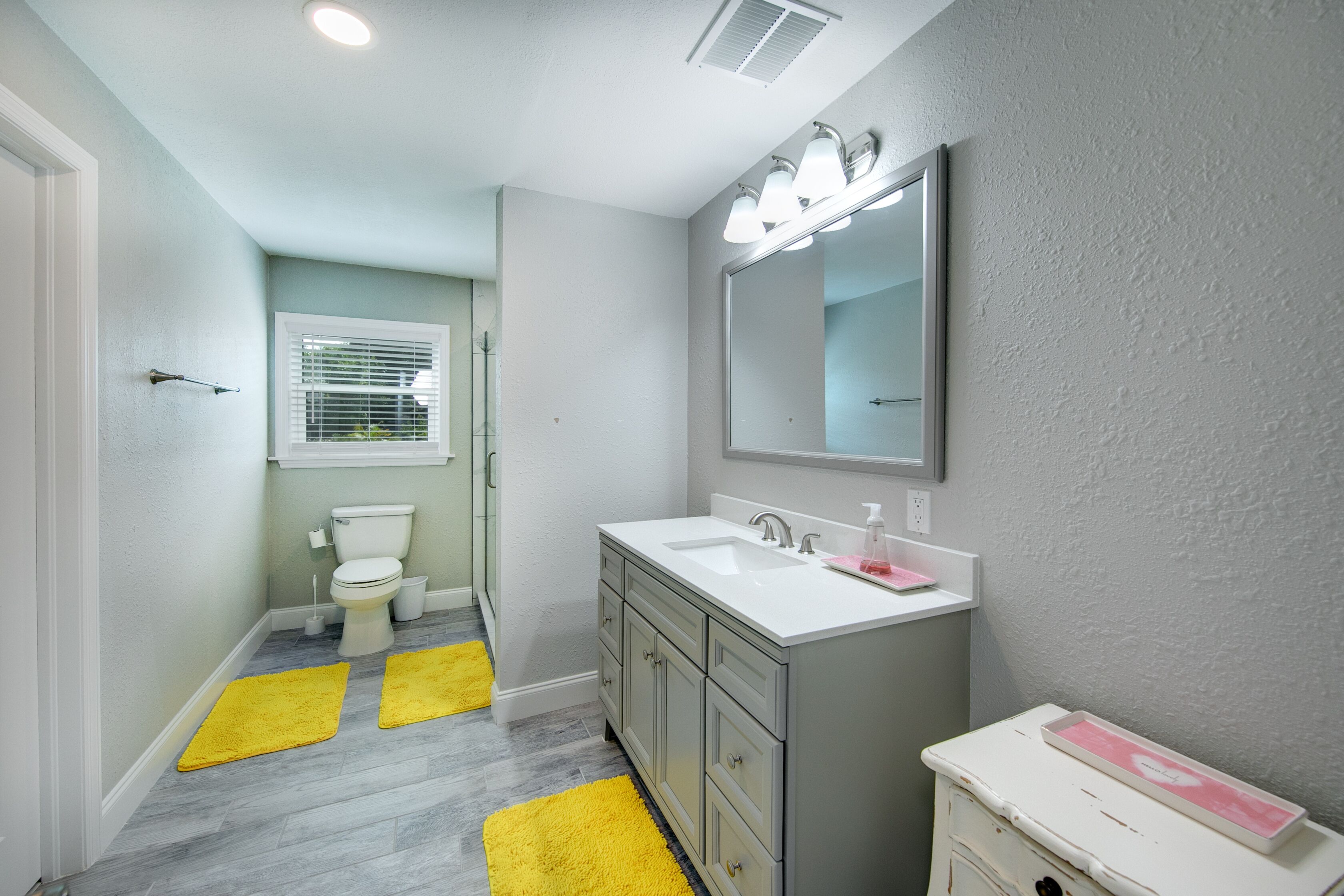 A bathroom with a white sink, a mirror, and a toilet. There are yellow rugs on the floor.