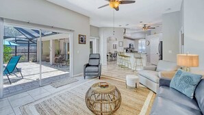 A spacious living area with a sofa, armchair, and coffee table. There is a ceiling fan, a kitchen area in the background, and a sliding glass door leading to an outdoor patio. - Haven Hideaway with Saltwater Pool 3br Sleeps 8 (Sarasota)