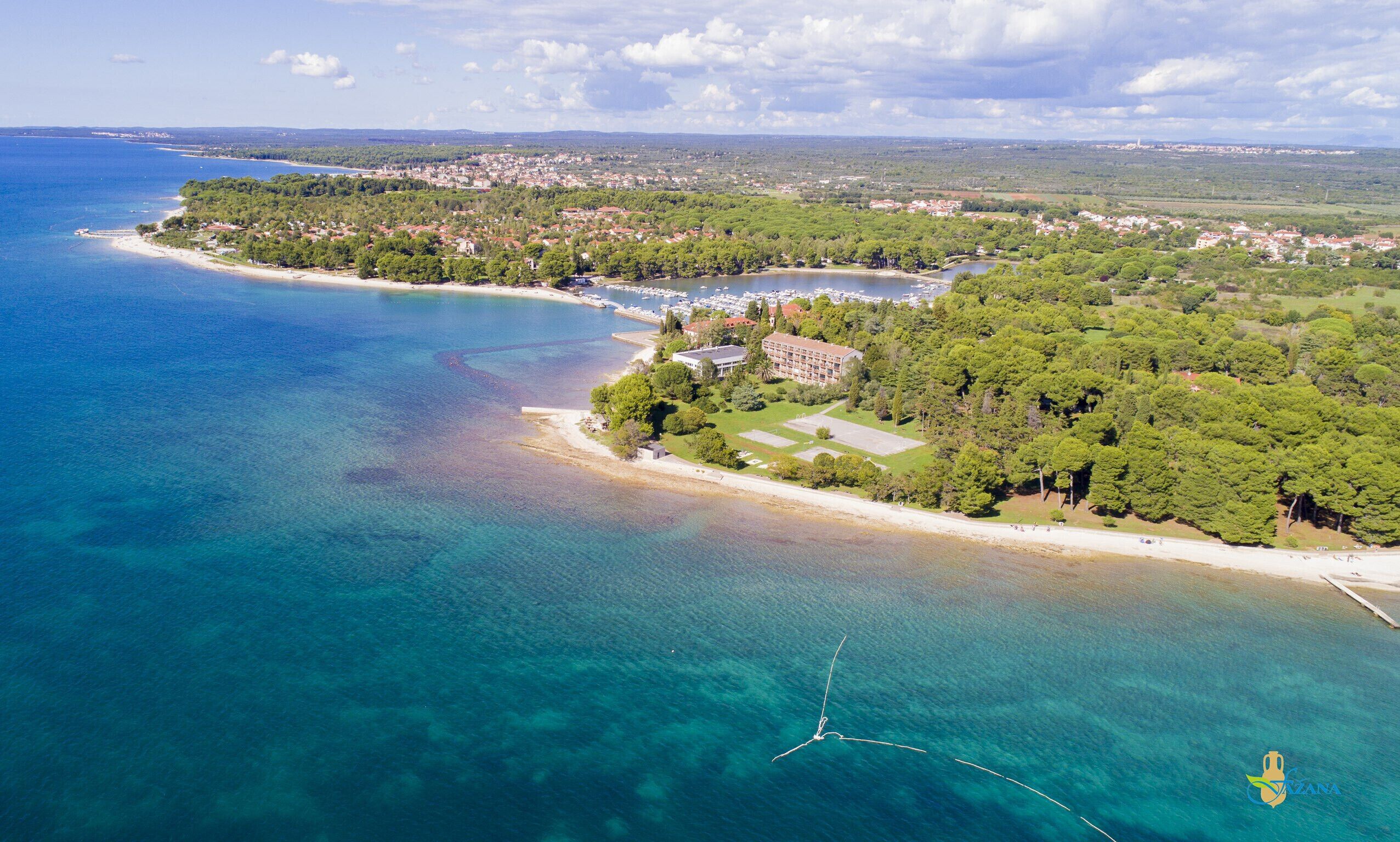 An aerial view of a coastal area with a beach, a marina, and a residential area with buildings surrounded by greenery.