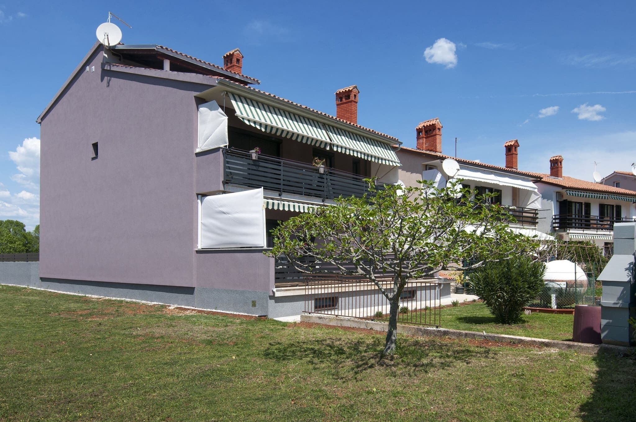 Una casa moderna de dos pisos con techo plano, antena parabólica y un pequeño árbol en el jardín delantero.