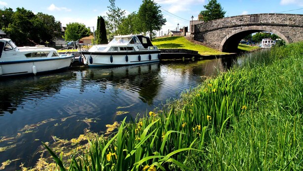 A canal with boats docked, a stone bridge, and lush greenery.