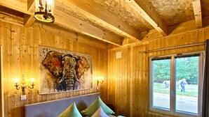 Ein Zimmer in einer Holzhütte mit einem Bett, Kissen, einem Elefantenbild und einem Fenster mit Blick auf einen Reiter und sein Pferd.
