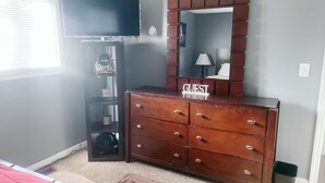 Une chambre à coucher avec une commode en bois, un miroir, une télévision et une fenêtre avec des stores.