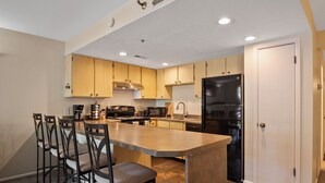 A modern kitchen with wooden cabinets, a central island with bar stools, a black refrigerator, and a microwave.