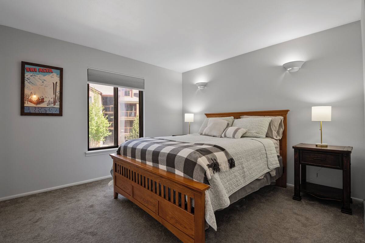 A bedroom with a wooden bed, two bedside lamps, a window with blinds, and a poster on the wall.