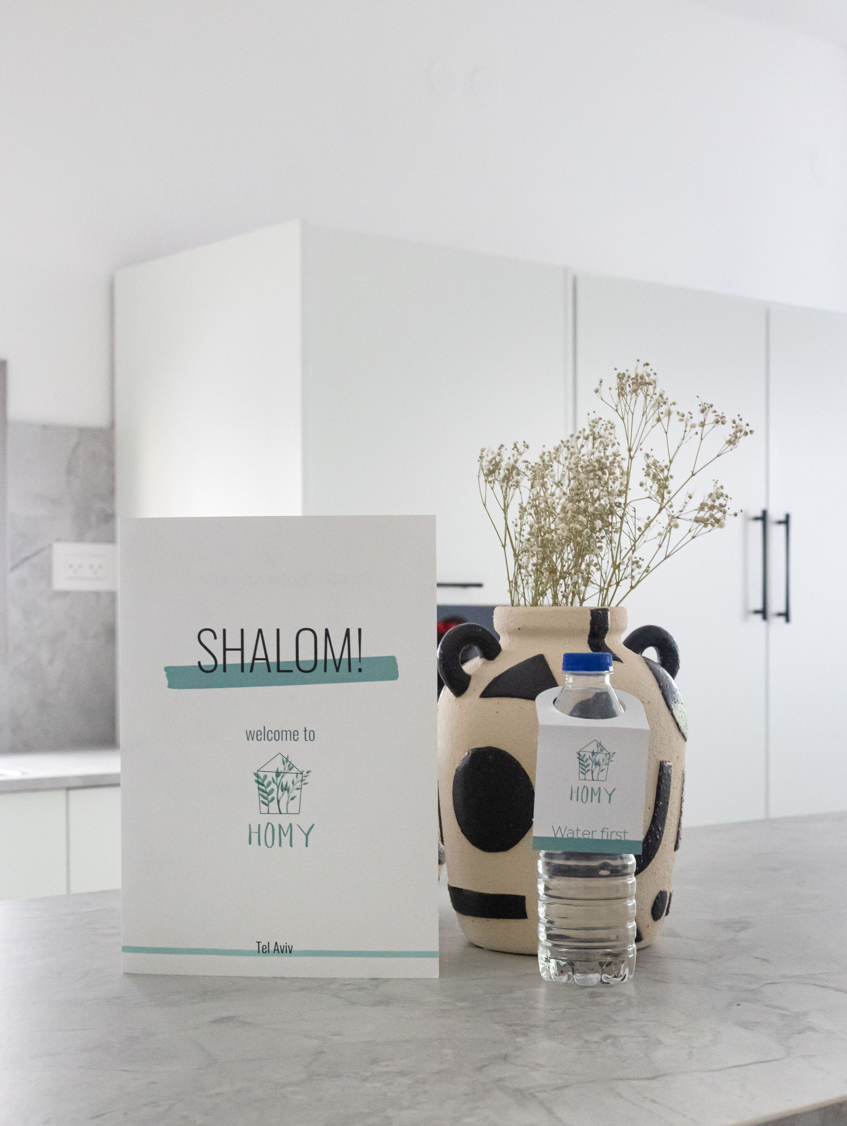 Une carte de bienvenue pour un hôtel nommé « SHALOMI », avec son logo, une bouteille d’eau et un vase rempli de fleurs, le tout posé sur un comptoir de cuisine.