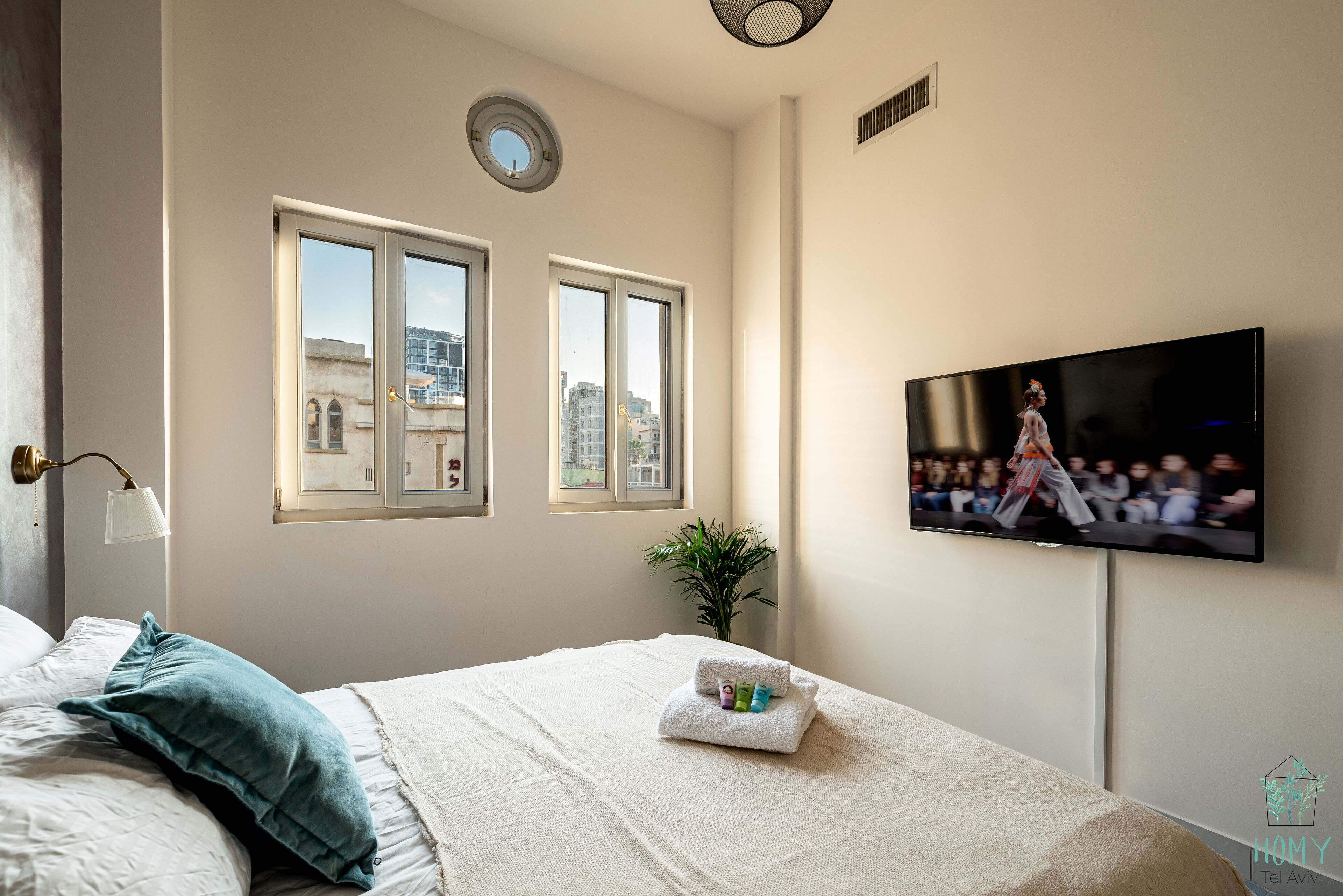 Una habitación de hotel con cama, televisor montado en la pared y ventana con vista a edificios.