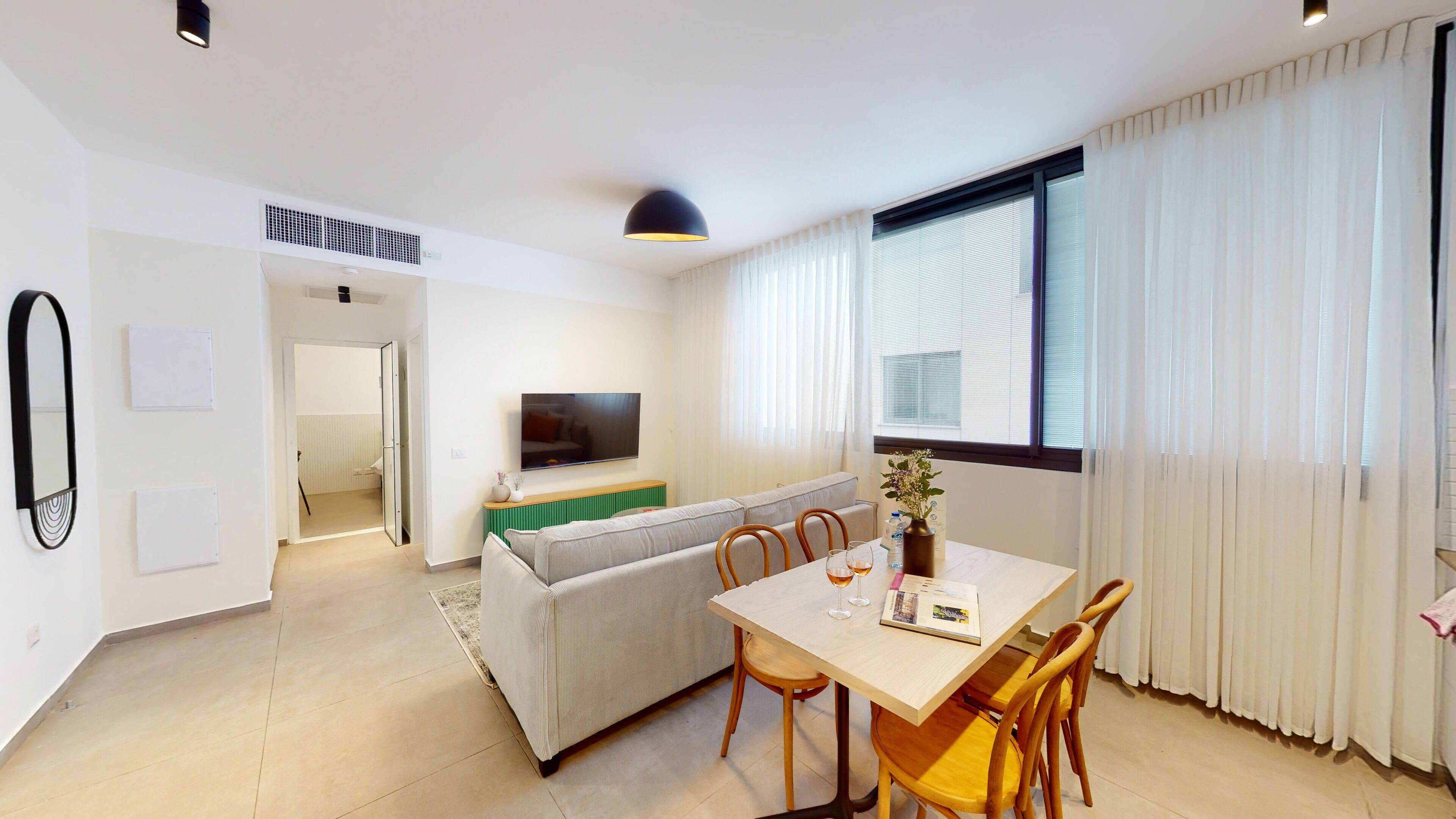 A modern living room with a sofa, dining table, and chairs. There is a flat-screen TV mounted on the wall, a window with curtains, and a ceiling light fixture.