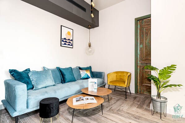 A modern living room with a light blue sofa, a black round ottoman, a wooden coffee table with magazines, a yellow armchair, and a potted plant.