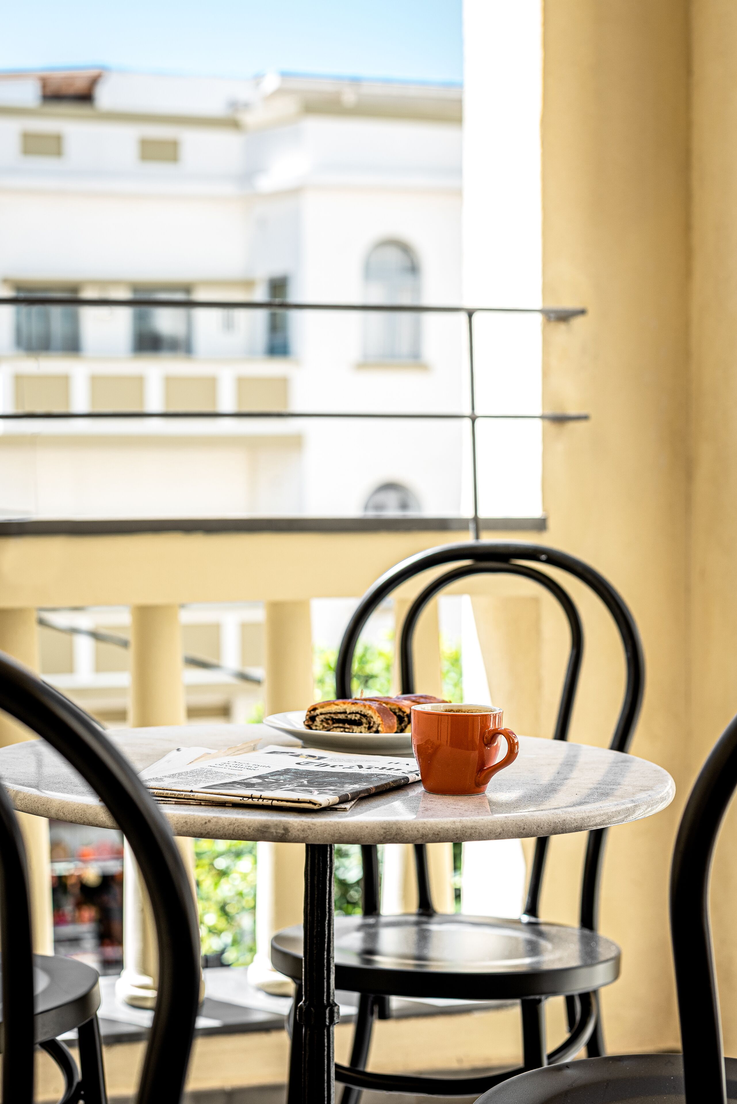 Una mesa redonda con un periódico, una taza y un pastel en un balcón con vistas a edificios.