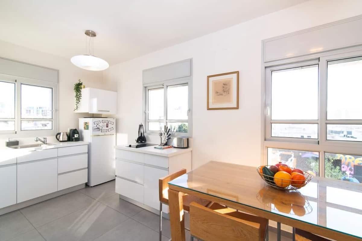 A modern kitchen with white cabinets, a glass dining table, and a bowl of fruit.