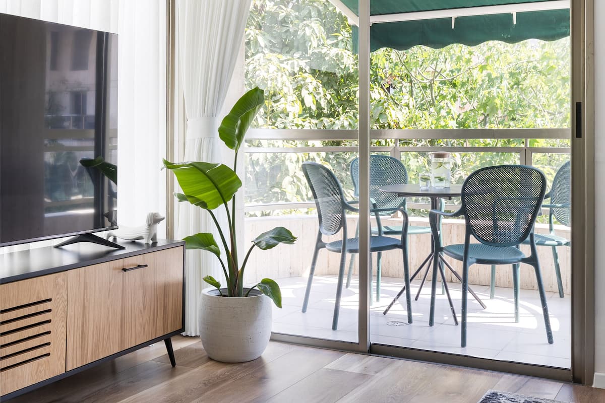 A modern living room with a flat-screen TV, wooden cabinet, potted plant, and a sliding glass door leading to an outdoor patio with chairs and a table.