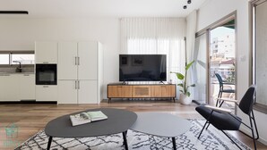 Een moderne woonkamer met een flatscreen televisie, een eethoek met een kleine tafel en stoelen, een keuken met witte kasten en een inbouwoven, en een groot raam met dunne gordijnen.