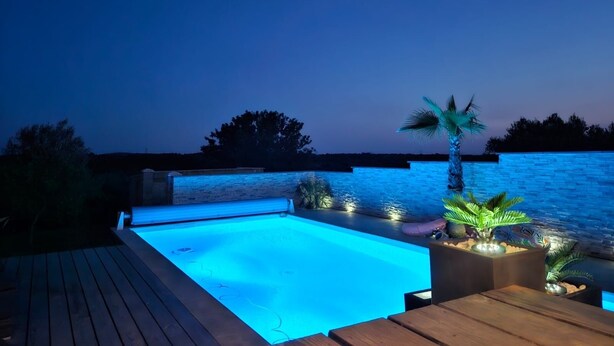 A swimming pool with a wooden deck and stone wall, surrounded by plants and palm trees, at dusk. - Magnificent villa Chloéline: Swimming pool and superb view (Pougnadoresse)