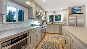 A modern kitchen with stainless steel appliances, light wood cabinets, and large windows.