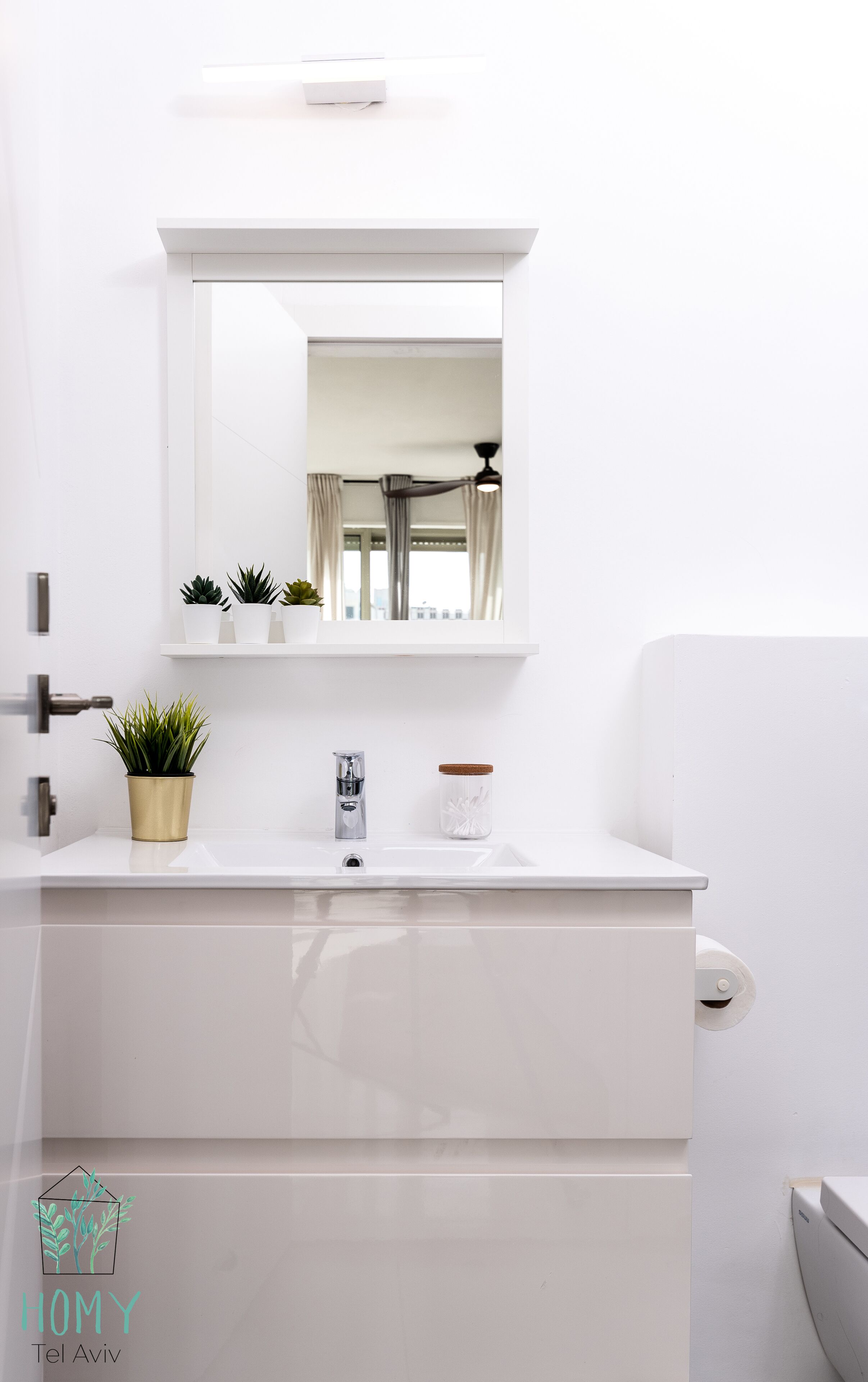 Un baño moderno con tocador blanco, espejo, grifo y retrete. Hay dos plantas en macetas sobre el tocador y un bote de bolas de algodón en la encimera.