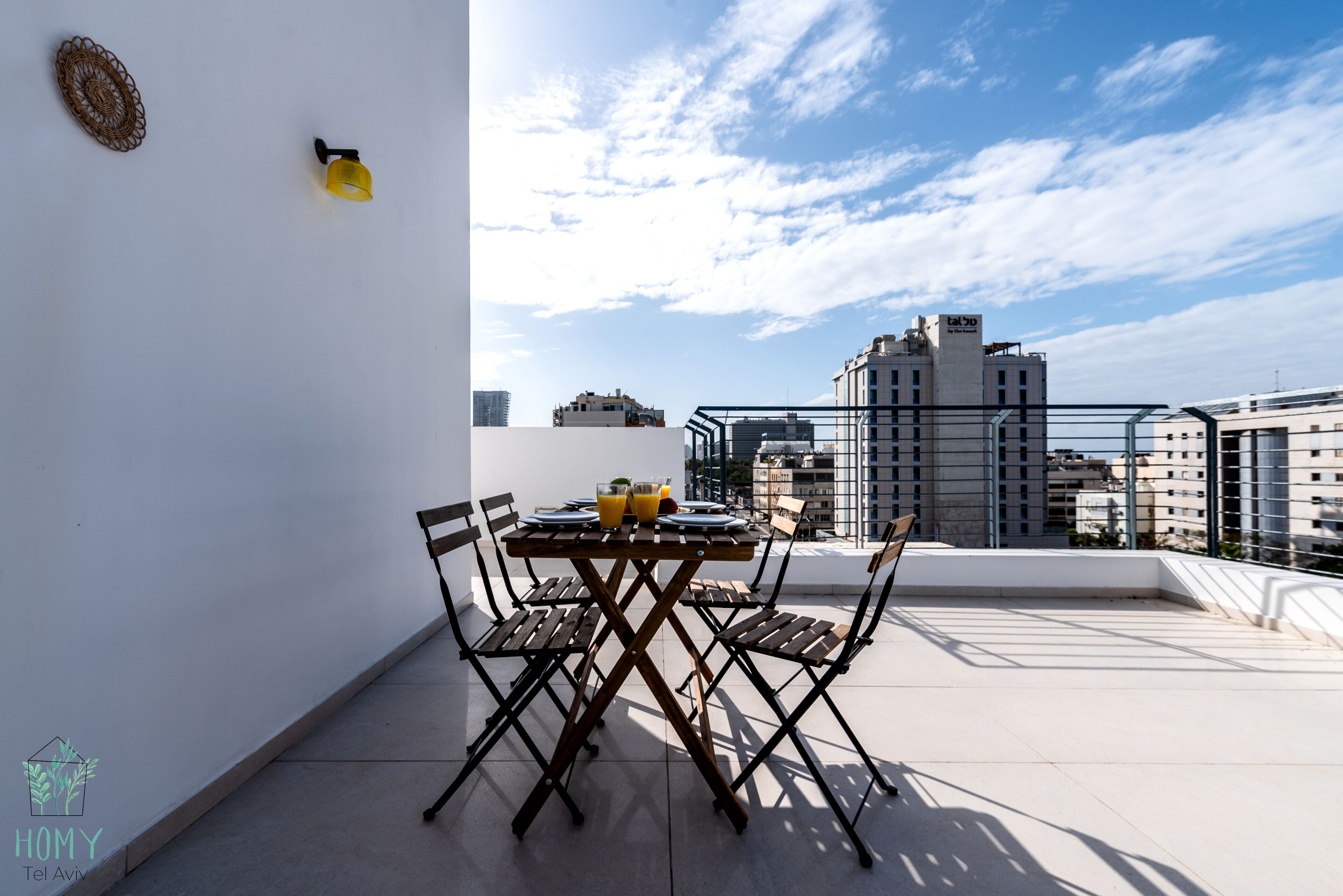 A rooftop terrace with a table set for two, two chairs, a yellow light fixture, and a view of the city skyline.