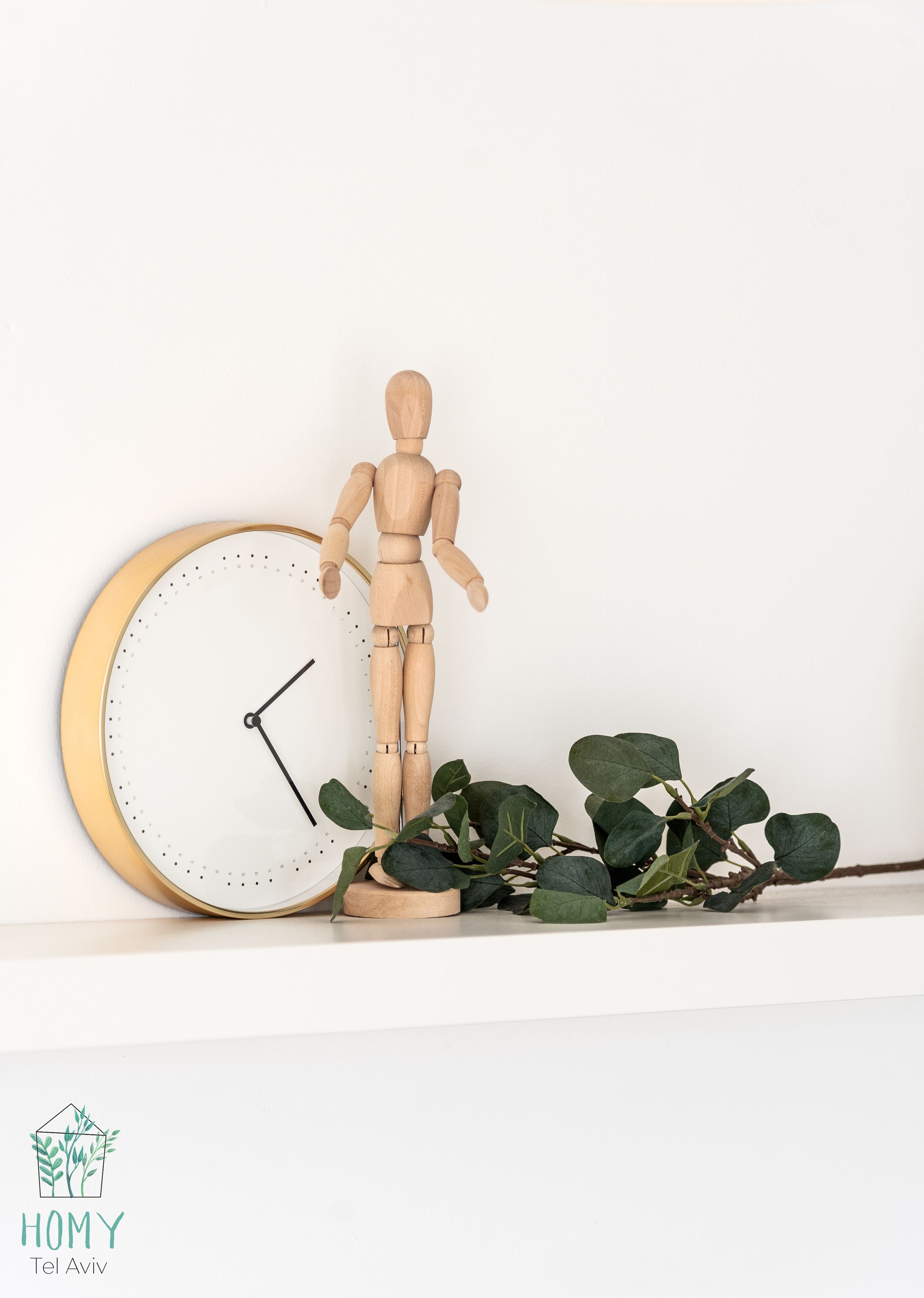 Eine Holzpuppe neben einer an der Wand befestigten Uhr mit goldener Rahmung, umgeben von grünem Grünwerk.