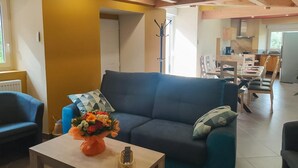 A modern living room with a dark blue sofa, a wooden coffee table with a vase of flowers, and a dining area in the background. - Lou Vachou - Rimeize (RIMEIZE)