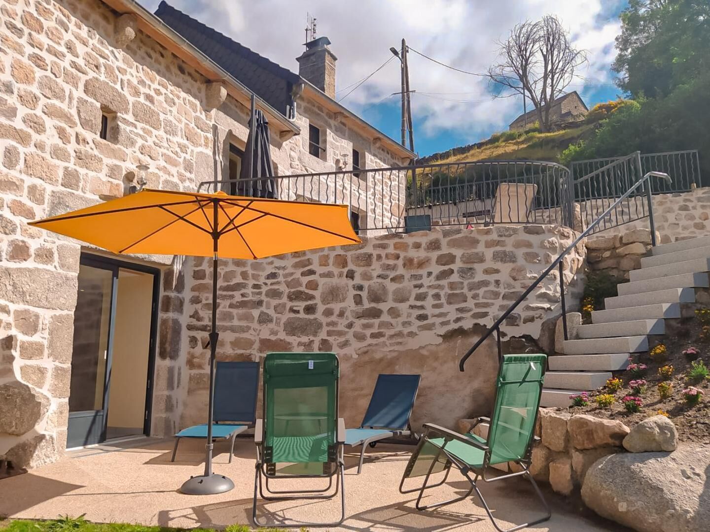 A stone building with a staircase leading to a higher level. There are outdoor chairs and an umbrella.