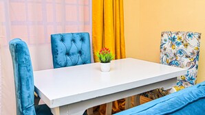 Zona pranzo con tavolo bianco, sedie imbottite blu, una piccola pianta in vaso con fiori rossi e un pannello decorativo con motivi floreali.