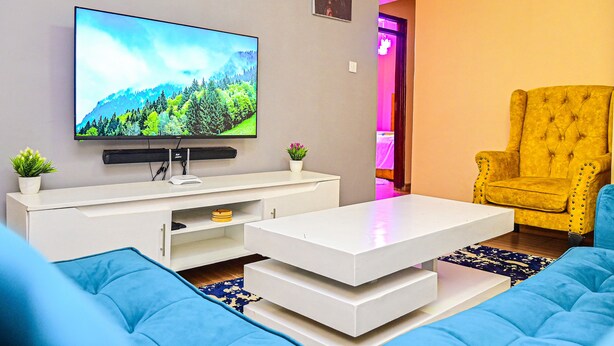 Un soggiorno moderno con una TV a schermo piatto, un mobile bianco per l'intrattenimento, un divano blu, una poltrona gialla e una scala che conduce a un piano superiore.