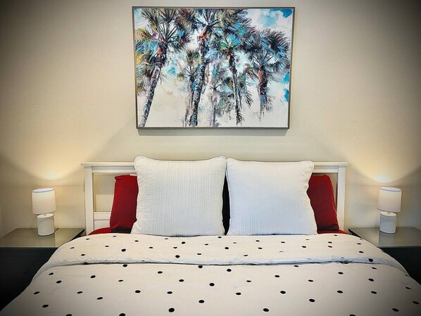 Una habitación de hotel con una cama, ropa de cama blanca, almohadas rojas y un cuadro de palmeras en la pared.