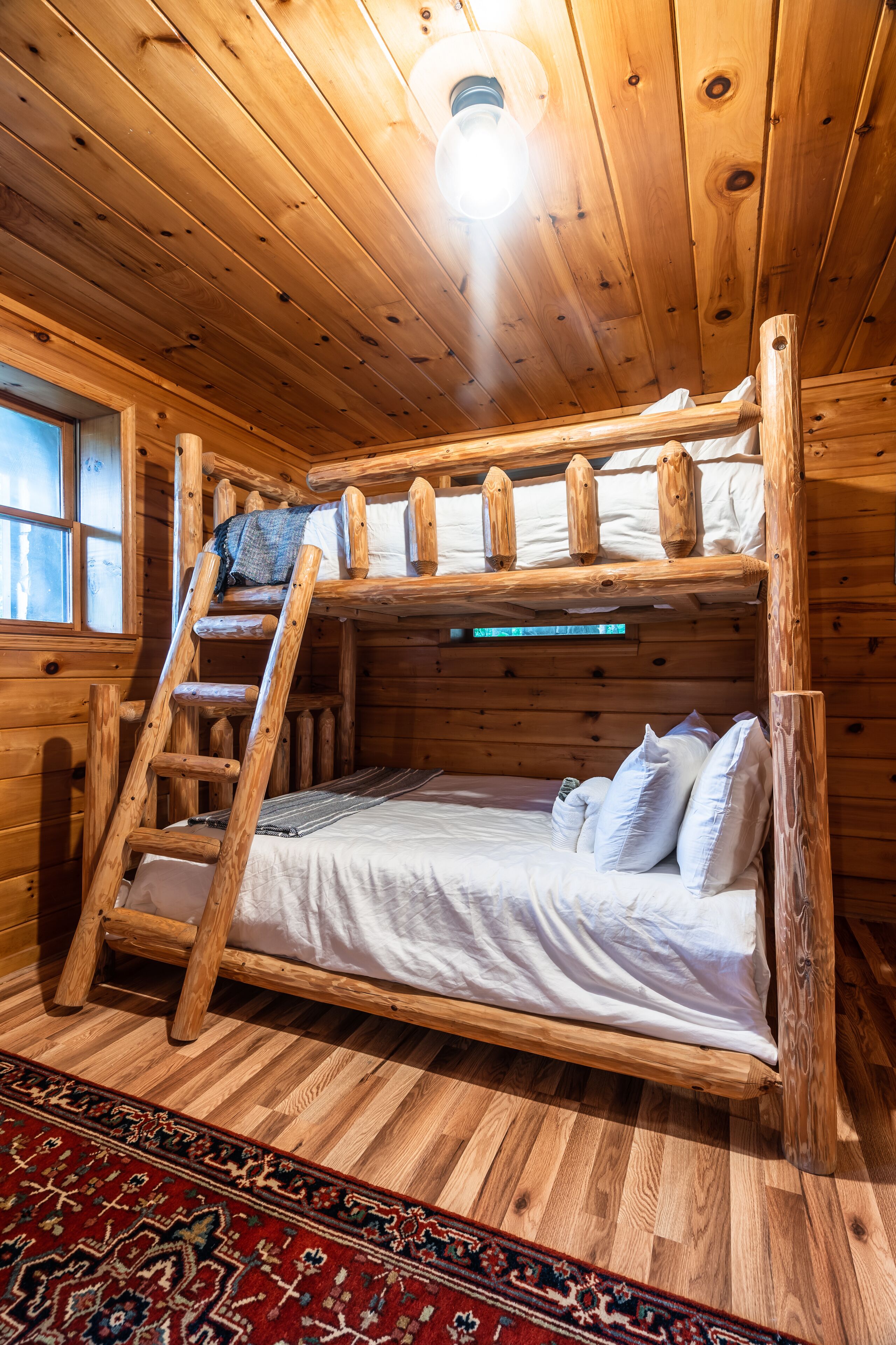 A wooden cabin room with a bunk bed, a ladder, a window, and a patterned rug.