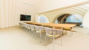Uma sala de conferências com uma mesa comprida e cadeiras, uma televisão na parede e vista para uma escadaria e uma estrutura azul através de um painel de vidro.
