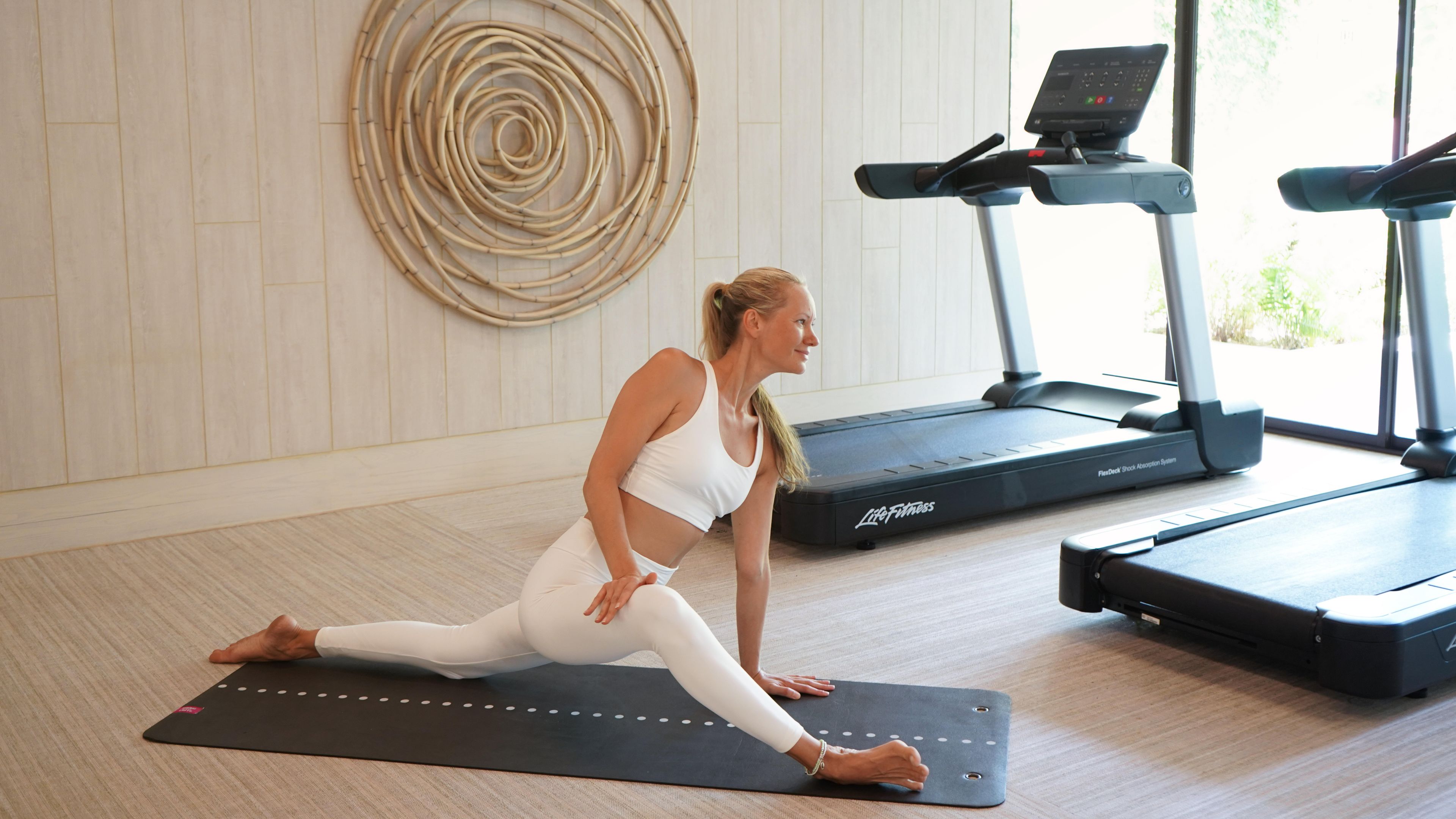 Una mujer realizando una postura de yoga sobre una colchoneta en una habitación con una cinta de correr y un adorno circular en la pared.