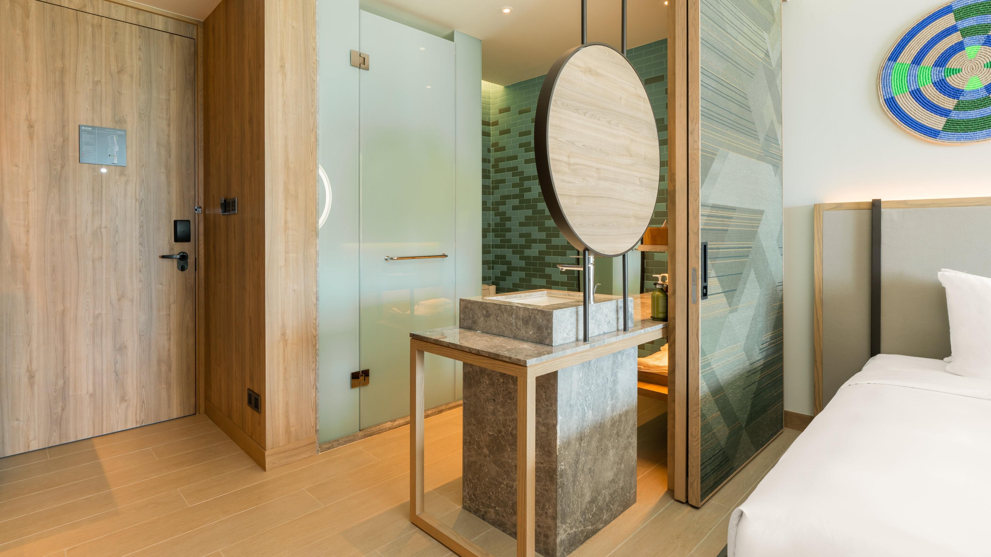 Una habitación de hotel moderna con un armario de madera, una zona de ducha con mampara de vidrio, un lavamanos de mármol y una cama con ropa de cama blanca.