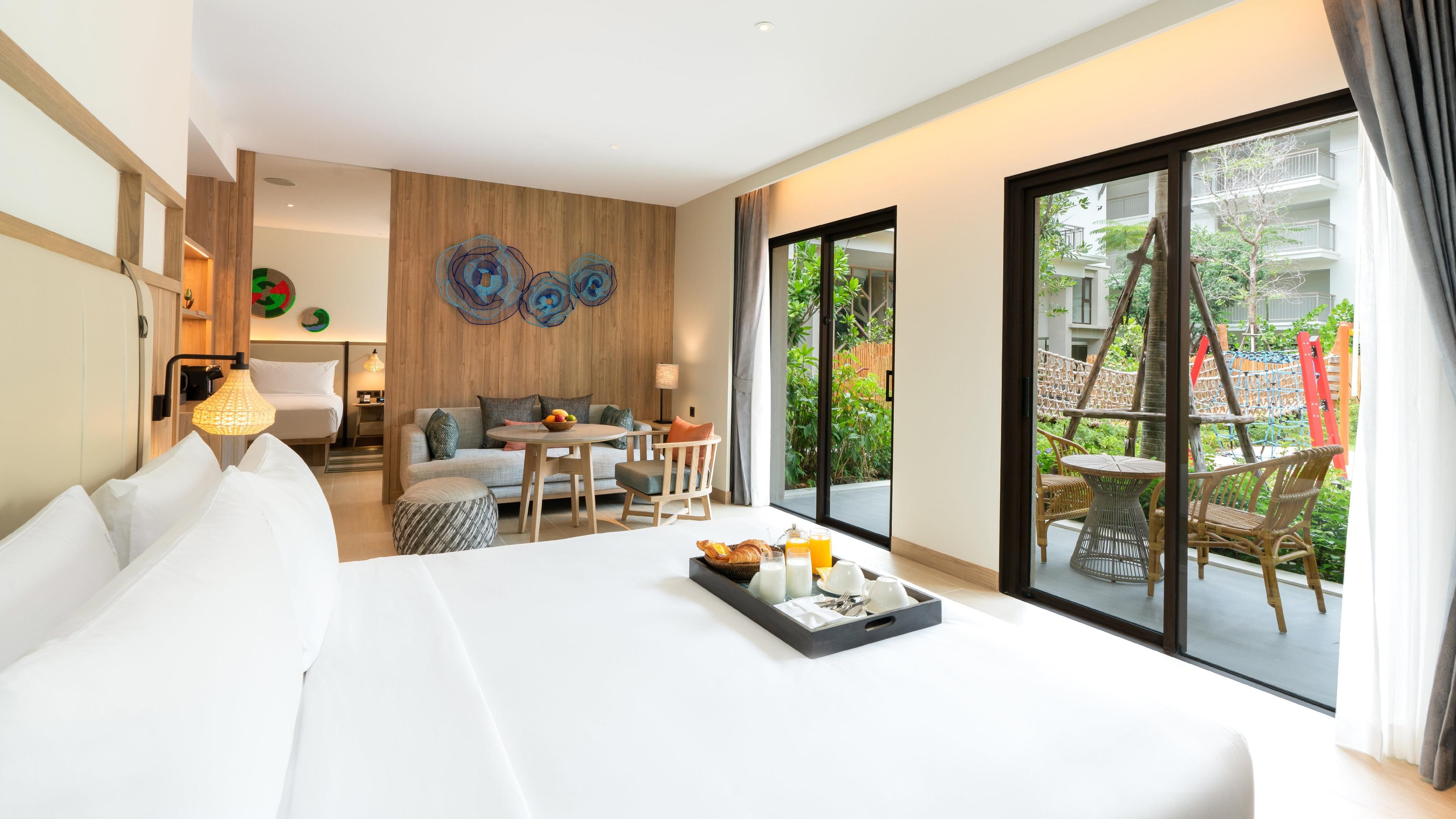 Habitación de hotel con cama, zona de estar con mesa y sillas, ventana grande con vistas a la vegetación y una pared de acento de madera.