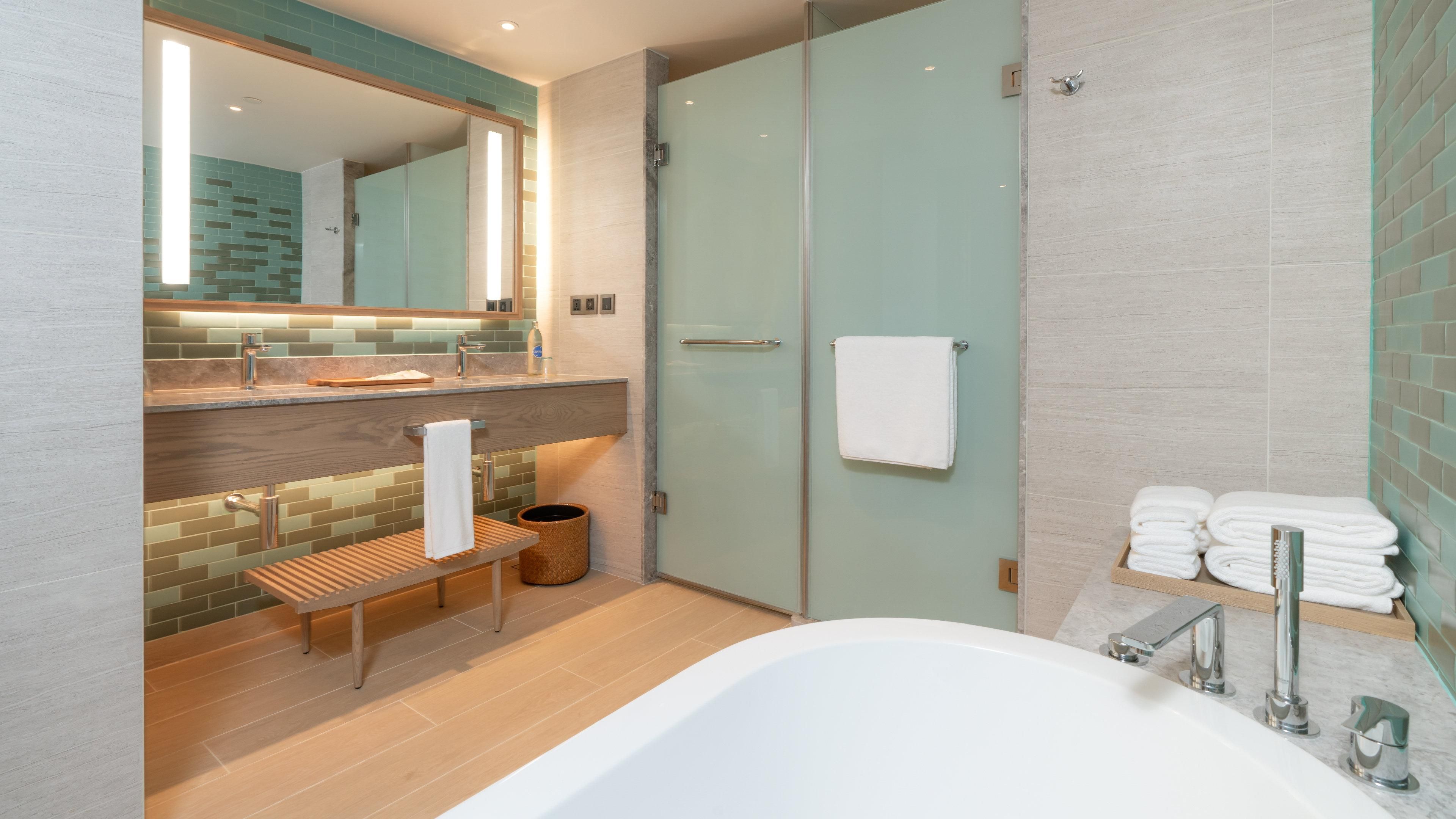 Un baño moderno con cabina de ducha de vidrio, un banco de madera, una bañera grande y un perchero con toallas dobladas con cuidado.