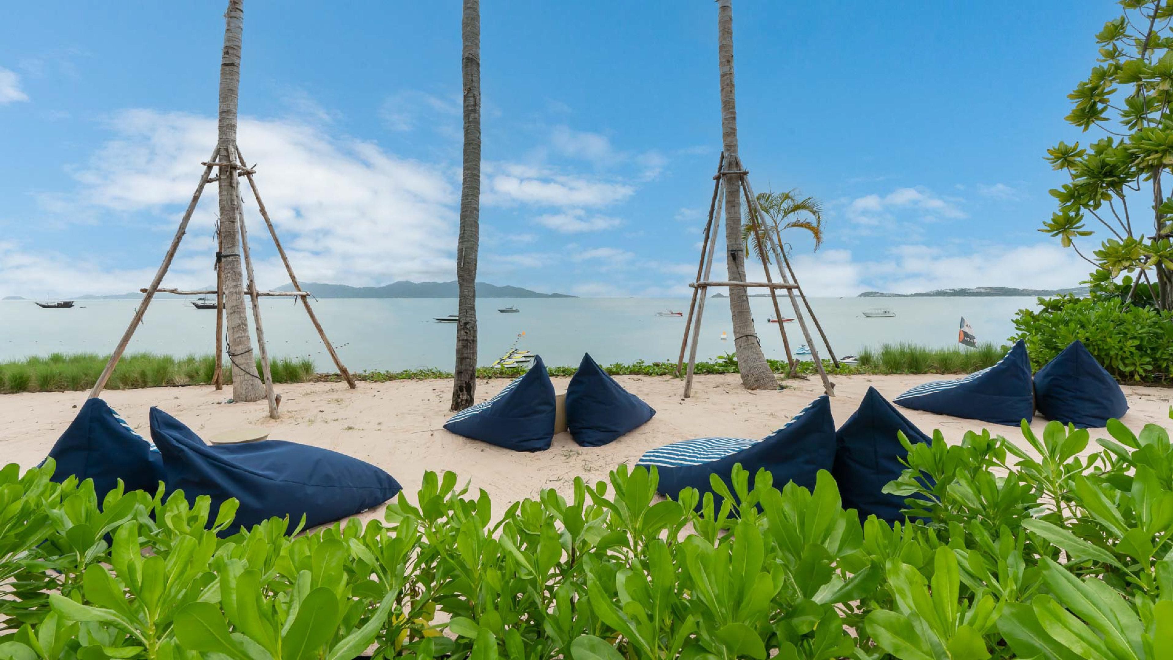Una playa con hamacas y pufs bajo palmeras, con vista a aguas tranquilas y barcos.