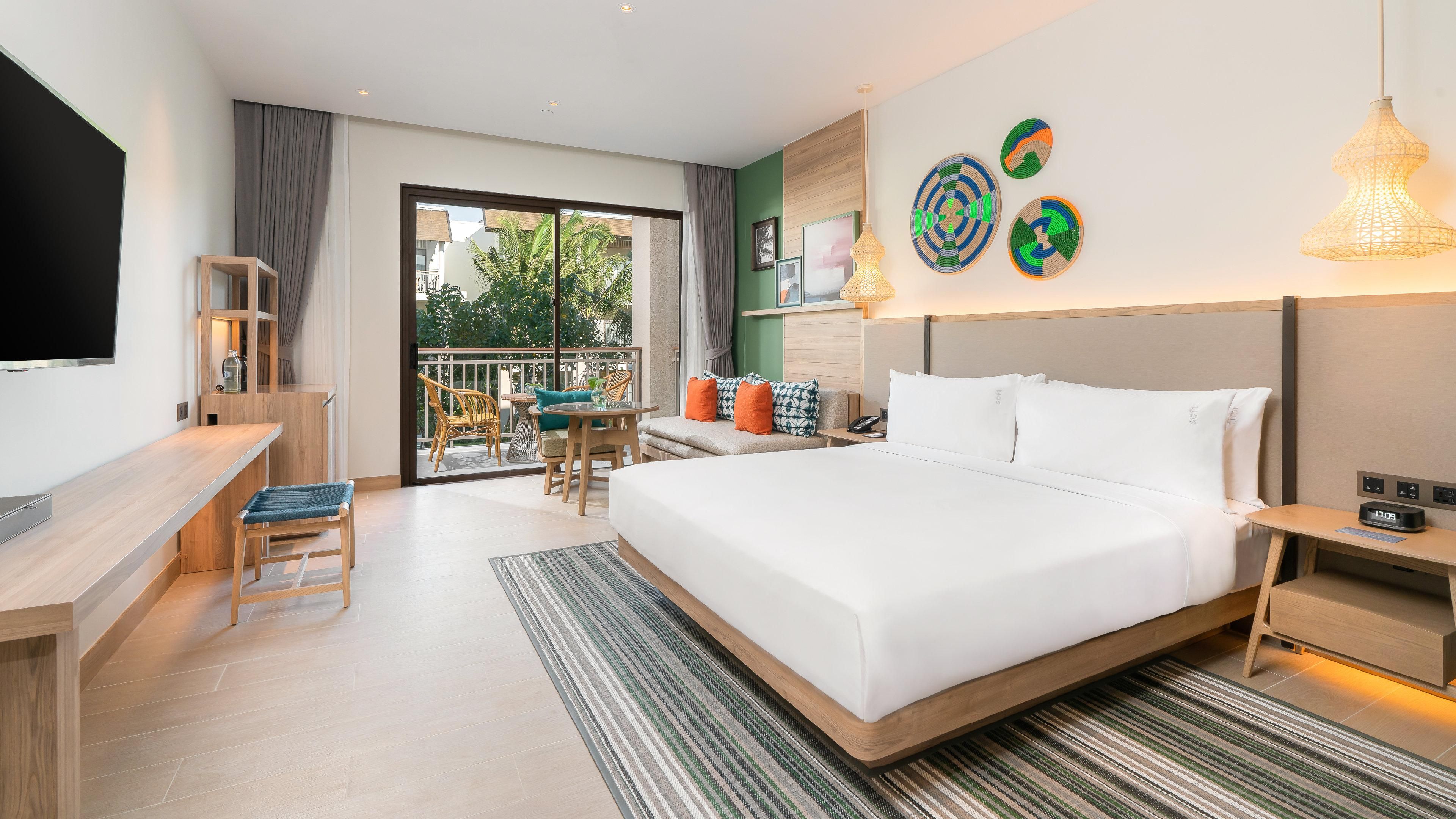 Una habitación de hotel moderna con una cama grande, un escritorio, una silla, un balcón con mobiliario exterior y obras de arte en las paredes.