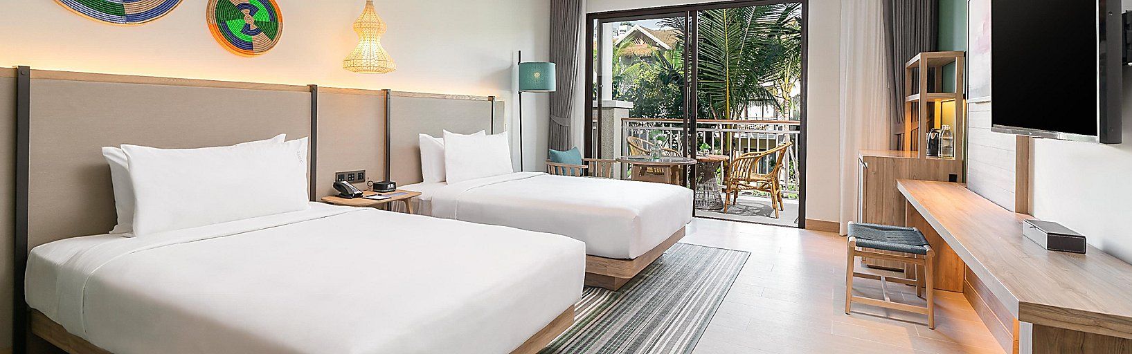 A hotel room with two beds, a desk, a TV, and a balcony with outdoor furniture.