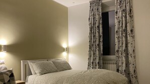 Un dormitorio con cama, mesita de noche, lámpara, ventana con cortinas florales y vistas al exterior.
