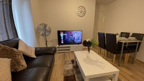 Una sala de estar moderna con un sofá de cuero negro, una mesa de centro blanca, un televisor de pantalla plana, un ventilador de pie y una zona de comedor con sillas negras y una mesa blanca.