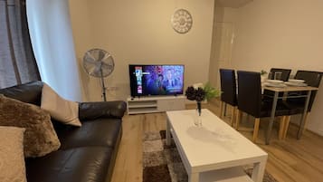 A modern living room with a black leather sofa, a white coffee table, a flat-screen TV, a standing fan, and a dining area with black chairs and a white table.