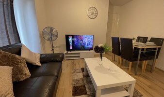 Uma sala de estar moderna com um sofá de couro preto, uma mesa de centro branca, uma TV de tela plana, um ventilador de pedestal e uma área de jantar com cadeiras pretas e uma mesa branca.