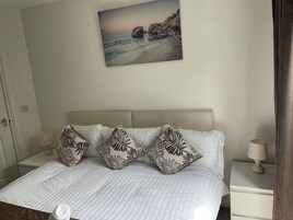 Quarto com cama, almofadas, mesa de cabeceira, abajur e um quadro com uma paisagem de praia.